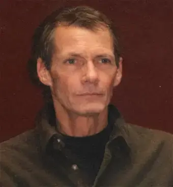 Man with brown hair, wearing brown dress shirt with black mock shirt under it.  Brown background.