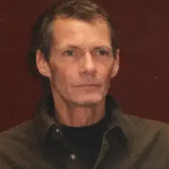 Man with brown hair, wearing brown dress shirt with black mock shirt under it.  Brown background.