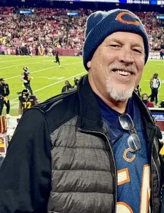 Man with gray goatee, wearing a toboggan, padded jacket standing in front of a game field  