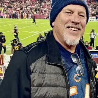 Man with gray goatee, wearing a toboggan, padded jacket standing in front of a game field  