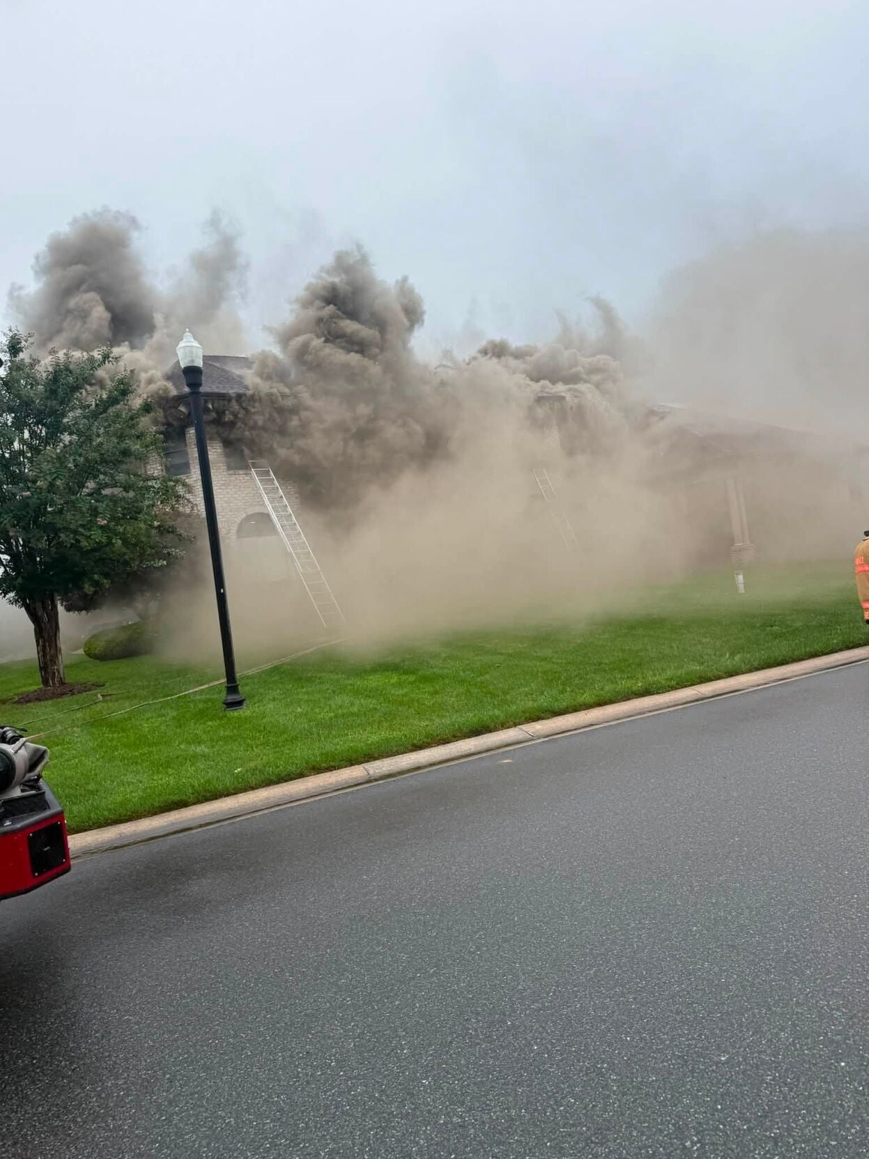 Large amounts of smoke billow from a two-story, brown brick home. A ladder sits against the front wall of the home up to a window, presumably from firefighters at the scene. The smoke obscures most of the home. The front yard has nicely cut, green grass. A black lamp post sits on the yard. Next to it is a small tree. The rear bumper of fire crew vehicle is visible on the edge of the left frame. The sky is blue.