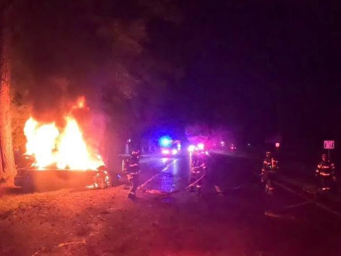 Firefighters stand in a roadway while responding to a crash scene off US 113 with a sedan fully engulfed by flames.