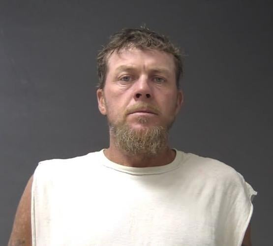 A white male with facial hair wears a white muscle tee in a mugshot.
