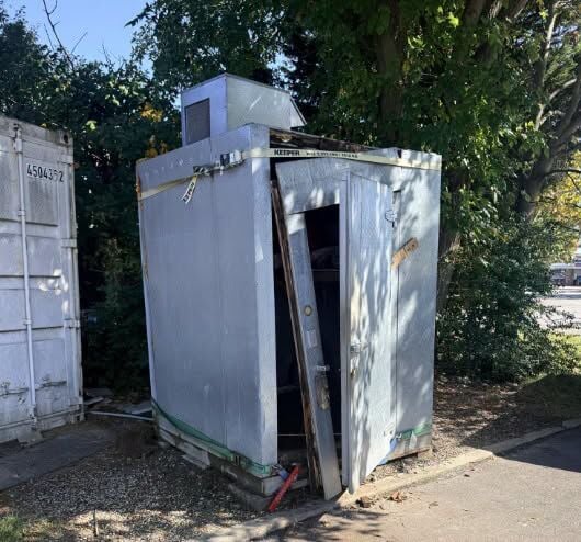 An outdoor walk-in cooler sits open with the door partially pried off.