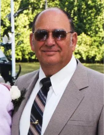 man with bald head, wearing sunglasses, white dress shirt, gray suit and trees in the background