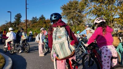 Elementary-age students sit on bikes with backpacks on a trail surrounded by trees. 