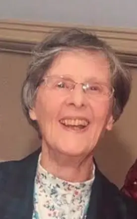 woman with short salt and pepper hair, wearing glasses, flower mock top with black jacket standing in front of a beige door. 