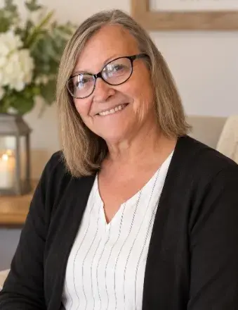women with shoulder length blond hair, wearing glasses, white v-neck top covered with black cardigan. Vase of flowers in back ground 