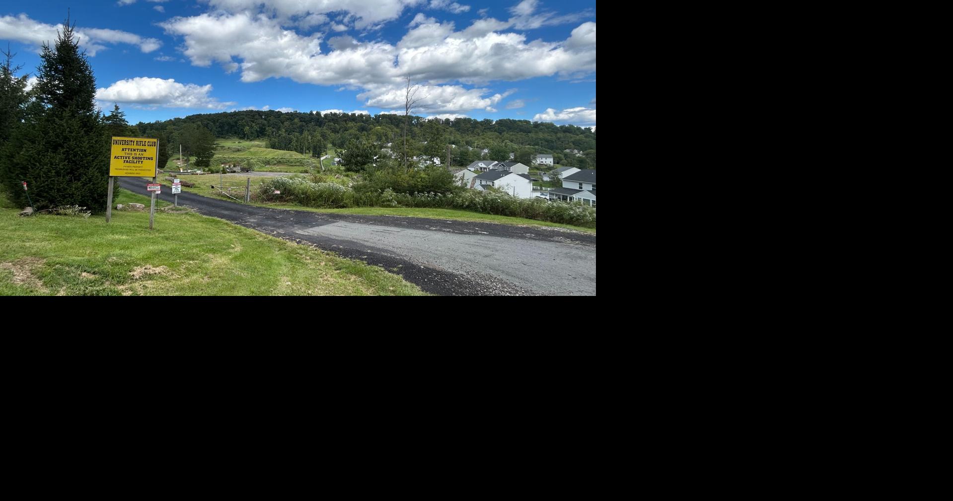 A yellow sign next to a driveway says “University Rifle Club. Attention this is an active shooting facility.” To the left of the road is a housing development and a hill in the background.