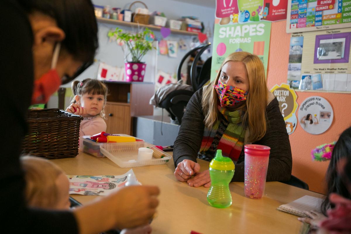 A masked woman sits at a table, with bottles and craft supplies covering the surface, while children in the foreground and on either side look on. 