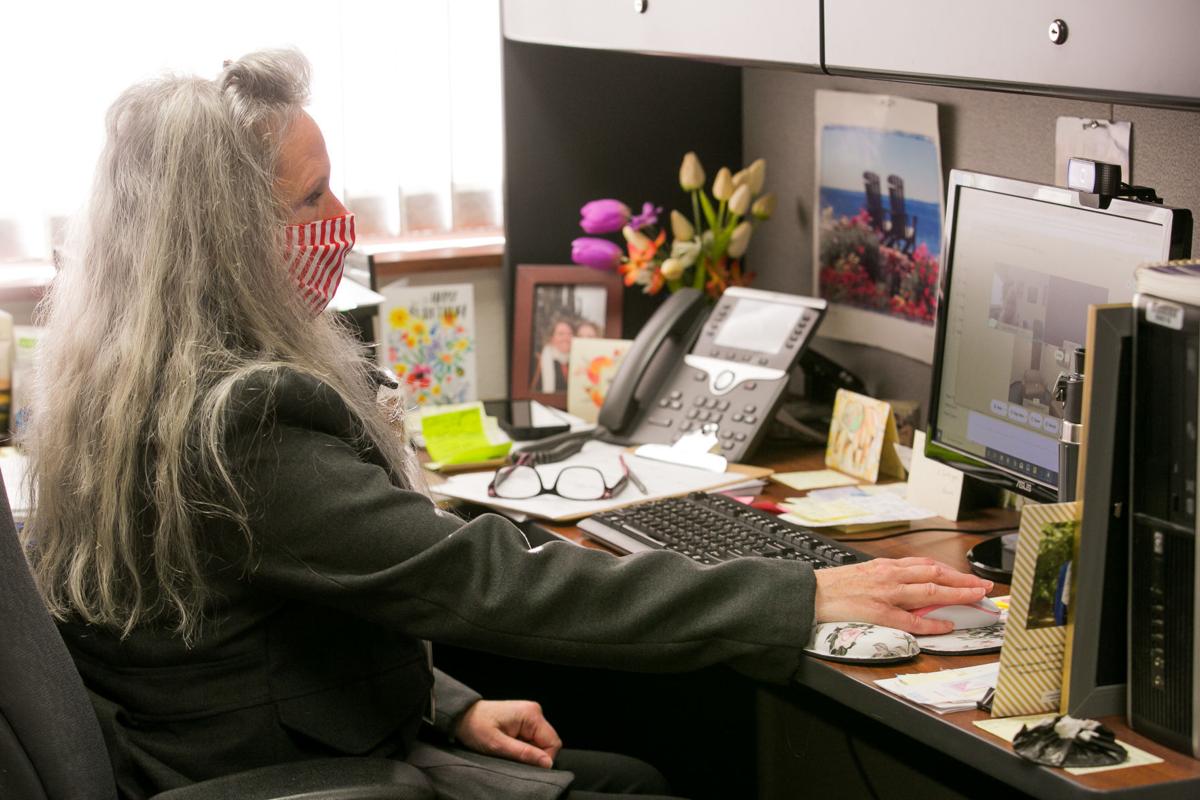 A person with long gray hair sits at a computer desk, with a bright window in the background. 