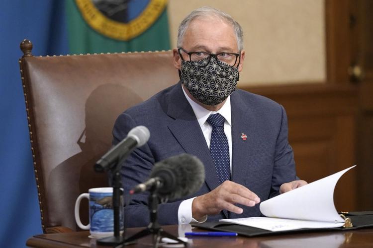 Gov. Jay Inslee sits at a wooden table in a tall, brown leather chair. A blue mug sits on the table next to a microphone, and he is wearing a dark blue suit. 